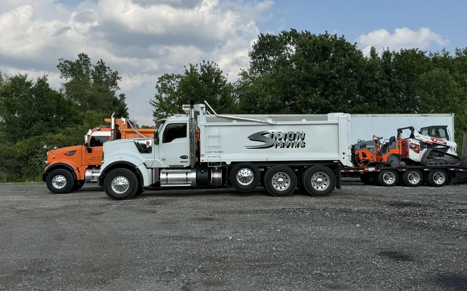 Simon Paving dump trucks and a trailer loaded with a paver, rollers and a skid steer