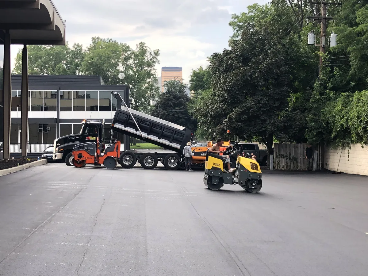 Rollers and a dump truck finishing a fresh asphalt lot beside a commercial building