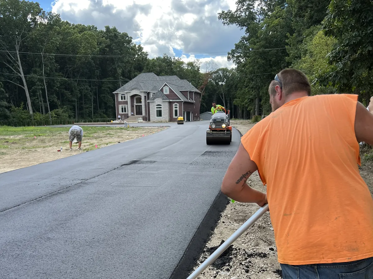 Crew member raking the edge of a freshly paved private drive leading up to a brick house